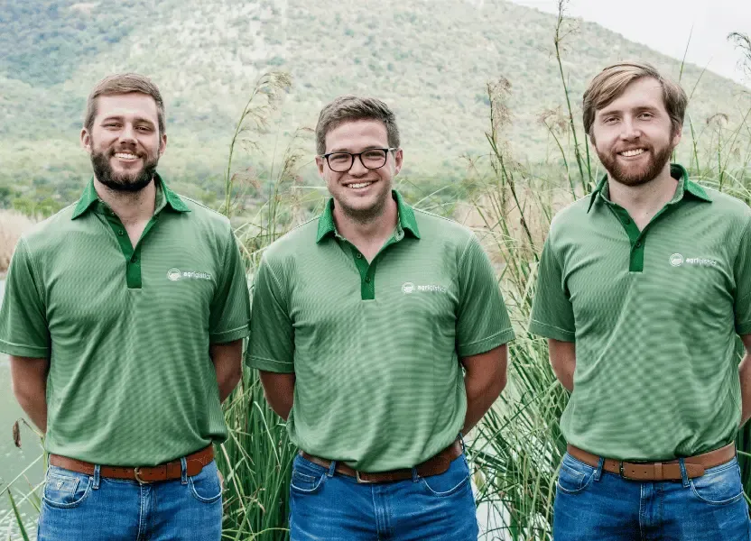 Three Agrigistics founders in green branded shirts standing outdoors in an agricultural field setting